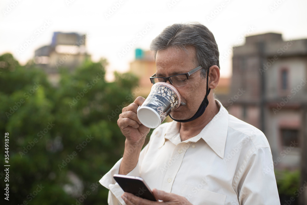 Portrait of an Indian old man with corona preventive mask watching his cell phone while drinking coffee on rooftop during afternoon in home isolation.Indian lifestyle, disease and home quarantine. Portrait of an Indian old man with corona preventive mask watching his cell phone while drinking coffee on rooftop during afternoon in home isolation.Indian lifestyle, disease and home quarantine.