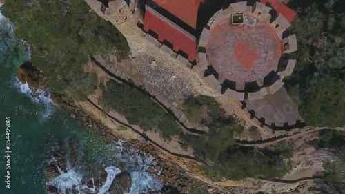 Aerial top view shot of waves splashing on rock formation by castle, drone flying over famous landmark on sunny day - Lloret de Mar, Spain photo