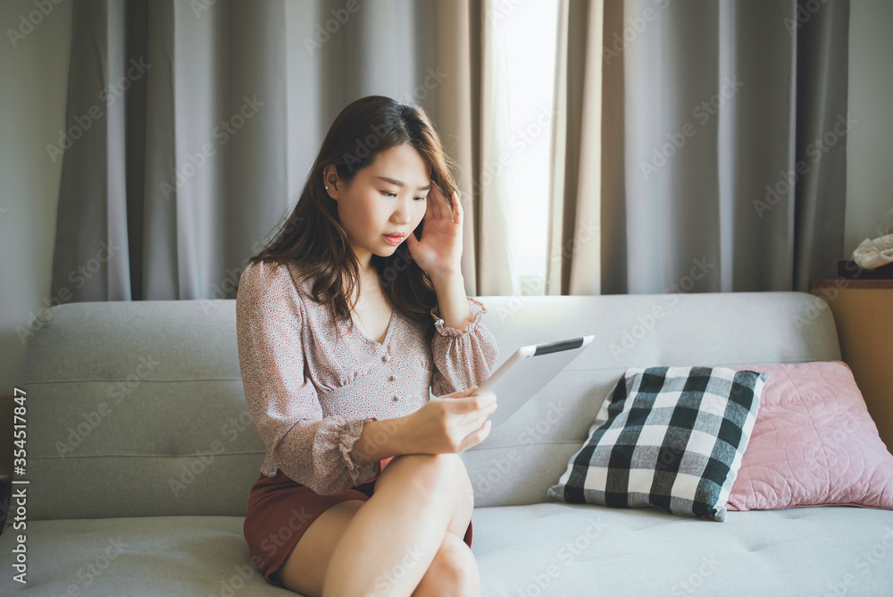 A beautiful asian woman is happy using tablet reading news article writing message sitting on the sofa at home. News people concept. A beautiful asian woman is happy using tablet reading news article writing message sitting on the sofa at home. News people concept.