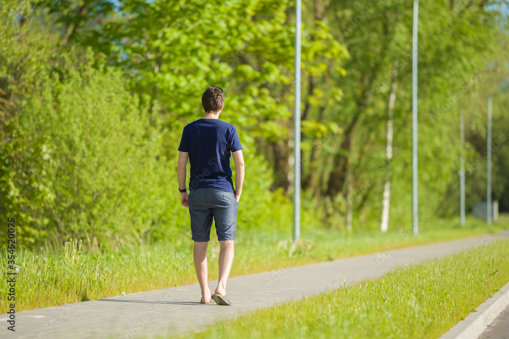 One young man in shorts and t-shirt slowly walking on long sidewalk at town green park. Warm, sunny spring day. Spending time alone in nature. Peaceful atmosphere. Back view. One young man in shorts and t-shirt slowly walking on long sidewalk at town green park. Warm, sunny spring day. Spending time alone in nature. Peaceful atmosphere. Back view.