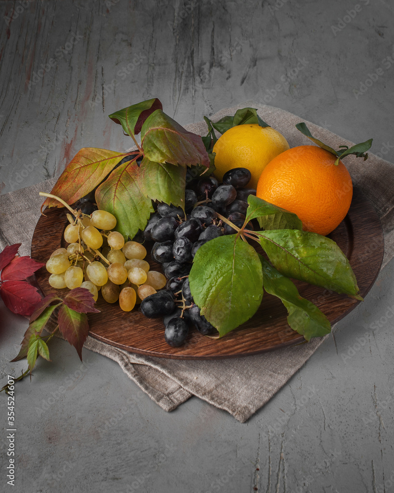 food photography of wooden plate with fresh fruits, grapes and oranges with leaves close-up on a gray texture background