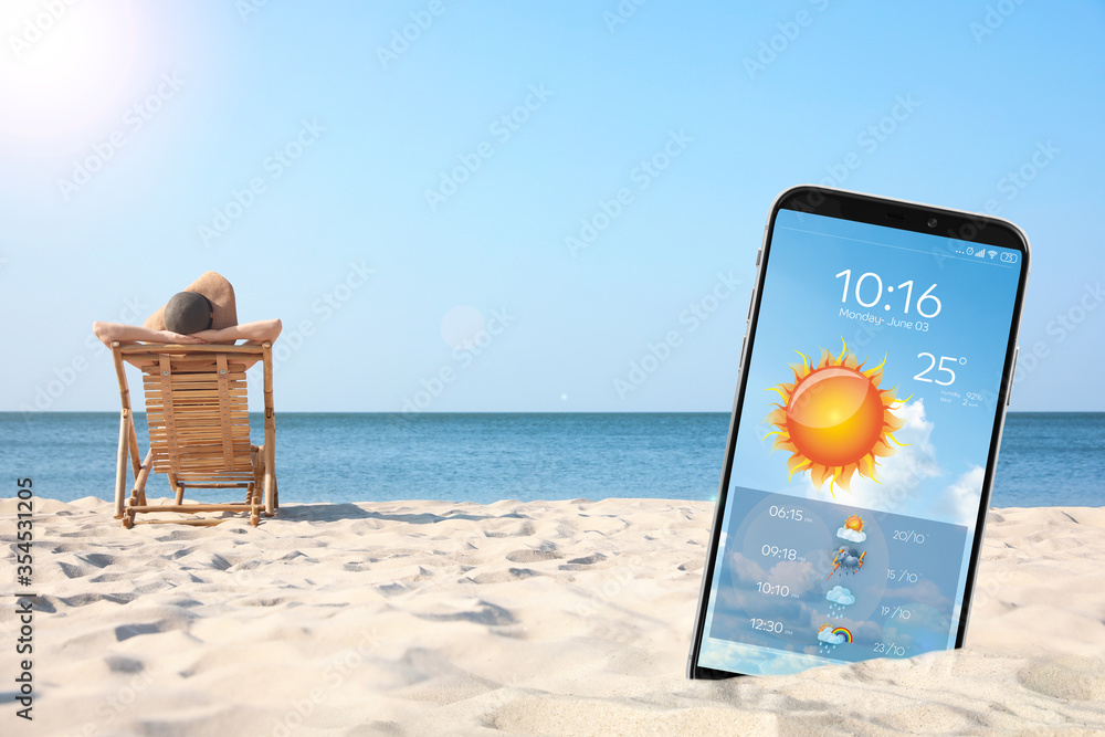 Young woman relaxing in deck chair on sandy beach and smartphone with open weather forecast app - obrazy, fototapety, plakaty 