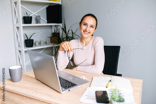 Cheerful young woman in office space. A woman with glasses looks at camera with smile sitting at the desk with a laptop