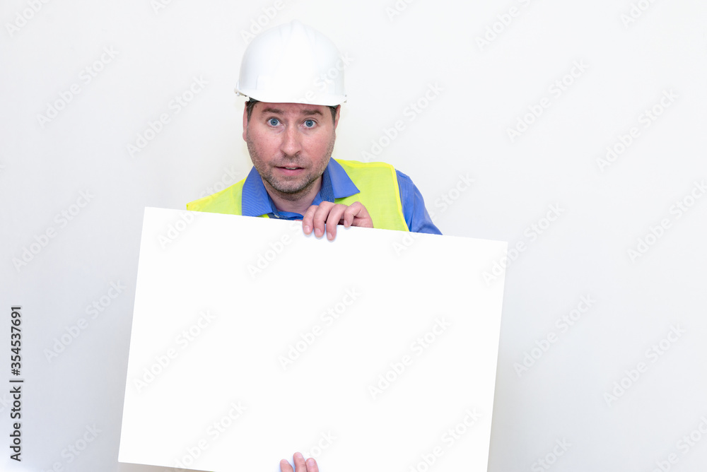 Close-up Engineer man wearing a helmet and reflector vest, standing and shows blank billboard. White studio wall. Close-up Engineer man wearing a helmet and reflector vest, standing and shows blank billboard. White studio wall.