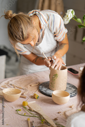Smiling beautiful girl in blue apron holding rolling pin and happily working with clay at pottery class studio. male potter master rolling up the clay on table with ceramic products.