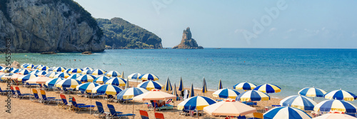 Agios Gordios beach, Corfu island, Greece. Panoramic view of the Greek beach - a long sandy sea shore with beach umbrellas and deck chairs, a blue sea and sky and unrecognizable people on the beach.