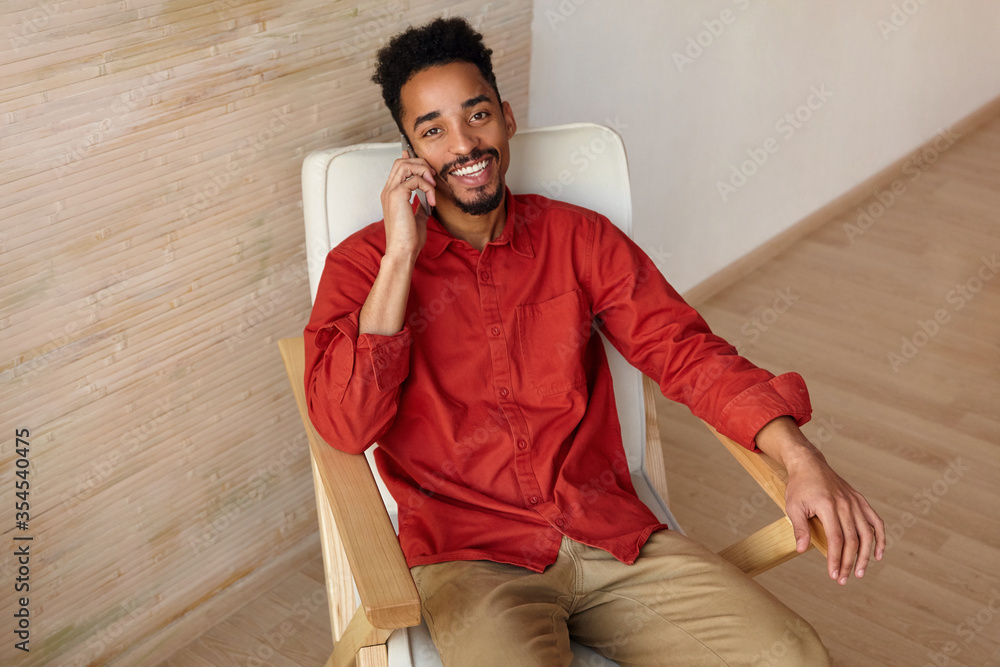 Happy young attractive brunette bearded dark skinned guy dressed in casual wear looking gladly at camera with pleasant smile while having phone talk, isolated over home interior