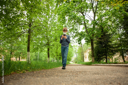 A masked teenager is walking in the park with a phone in his hand.