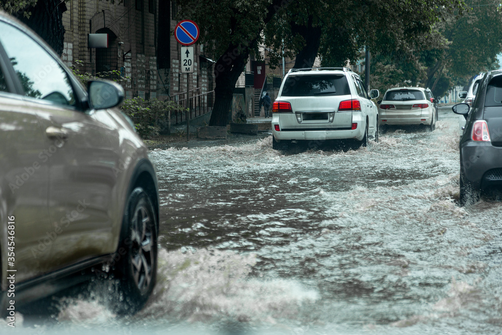 Вriving car on flooded road during flood caused by torrential rains. Cars float on water, flooding streets. Splash on car. Flooded city road with large puddle. Flooding after heavy rains at city Вriving car on flooded road during flood caused by torrential rains. Cars float on water, flooding streets. Splash on car. Flooded city road with large puddle. Flooding after heavy rains at city