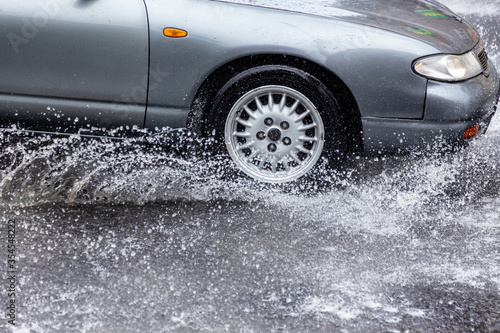 Вriving car on flooded road during flood caused by torrential rains. Cars float on water, flooding streets. Splash on car. Flooded city road with large puddle.  Flooding after heavy rains at city © Aleksandr Lesik