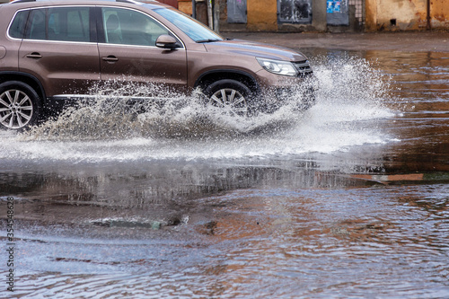 Вriving car on flooded road during flood caused by torrential rains. Cars float on water, flooding streets. Splash on car. Flooded city road with large puddle. Flooding after heavy rains at city