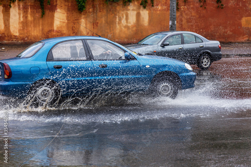 Вriving car on flooded road during flood caused by torrential rains. Cars float on water, flooding streets. Splash on car. Flooded city road with large puddle. Flooding after heavy rains at city