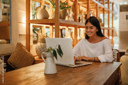  Smiling young woman in smart casual wear working on laptop while sitting near window in creative office or cafe. Young caucasian brunette woman using laptop computer in cafe
