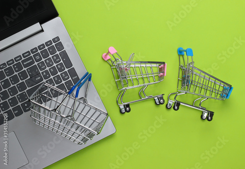 Online shopping. Laptop with supermarket trolleys, basket on green background. Top view. Flat lay