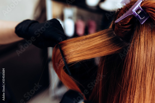 Hairdresser straightens redhead hair of woman with hair straightener tool in hair salon.