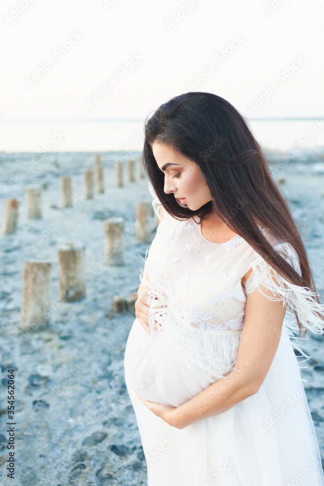 Beautiful young pregnant woman with long brown hair in white dress on the beach touching her belly with love and care on the background of the rocks and sea. Blur sea background. Maternity concept Beautiful young pregnant woman with long brown hair in white dress on the beach touching her belly with love and care on the background of the rocks and sea. Blur sea background. Maternity concept