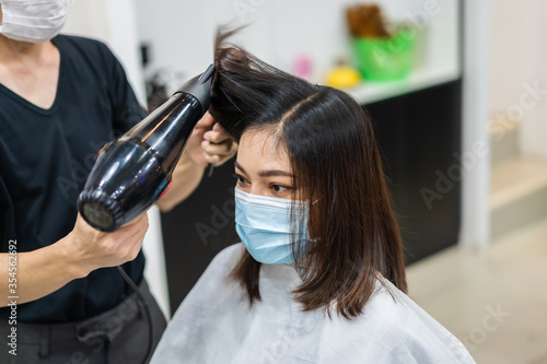 hairdresser using hair dryer and comb to the hair of woman, people must be wering medical mask to protection coronavirus (covid-19) pandemic. new normal concepts