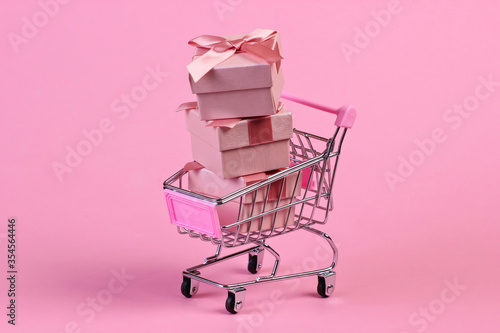 Holiday shopping. Mini supermarket trolley with gift boxes on pink pastel background