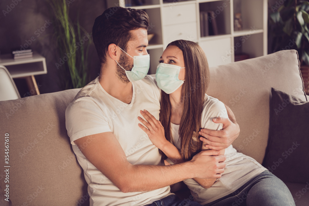 Photo of adorable young couple lady guy spend quarantine together stay home hugging look eyes happy together emotions sitting cosy sofa divan wear facial medical mask living room indoors