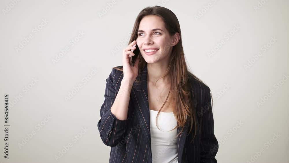 A smiling cheerful young woman wearing casual clothes is talking on the phone standing isolated over white background in studio A smiling cheerful young woman wearing casual clothes is talking on the phone standing isolated over white background in studio