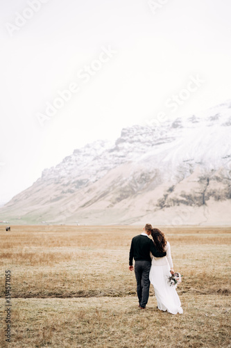 The wedding couple goes into the distance. The groom hugs the bride. Field of dry grass with moss, against the backdrop of a snowy mountain. Destination Iceland wedding.  © Nadtochiy