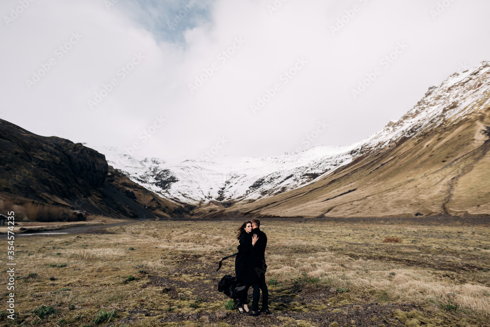 Destination Iceland wedding. Wedding couple on a background of snowy mountains. The bride and groom in black coats are hugging in a field of moss and yellow grass.
