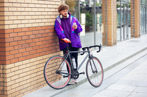 Fototapeta Naklejka Na Ścianę i Meble -  Handsome young man using mobile phone and headphones while standing near his bicycle. Listen to music, scrolling news, chatting, talking with friends on the go. All the services in device anywhere.
