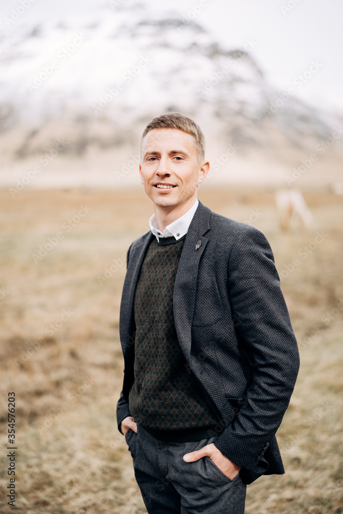 Destination Iceland wedding. Close-up man portrait. A guy in a sweater in a rhombus, a black jacket, stands in field of dry grass, holding his hands in his pockets, against backdrop of snowy mountain