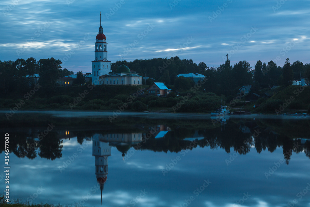 Idyllic Russian landscape in Totma at white night: orthodox church, rural houses and lonely ship on the bank of the Sukhona River with the reflection in water