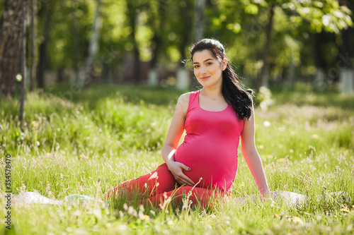 Attractive young pregnant woman doing yoga exercises outdoors in the park. Expecting female having healthy and active lifestyle.
