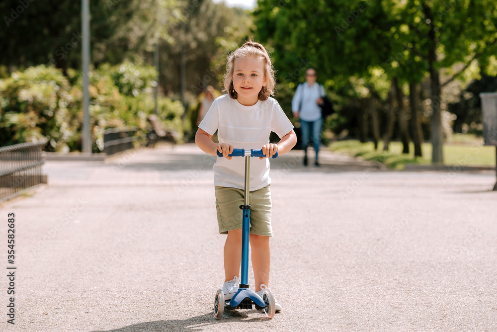 Fototapeta premium Boy learn to ride scooter in a park on sunny summer day. Preschooler boy riding a roller. Kids play outdoors with scooters.