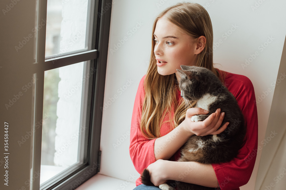 young woman holding cute cat while looking at window young woman holding cute cat while looking at window
