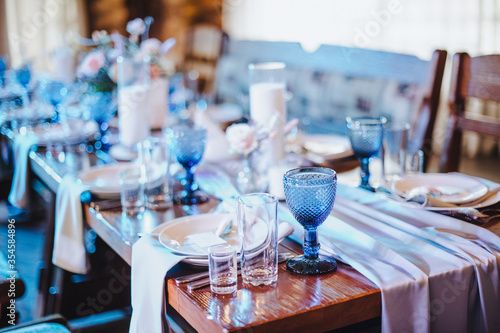 Rustic. The table is covered with a white tablecloth, served. Plates, cutlery, napkin, blue glasses, seating cards, a sprig of greens, candles in glass vases, blue light, dried flowers.