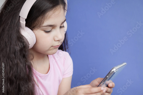 Cute child girl in headphones is using a smartphone, looking at camera and smiling on bright background. Listening music. Online talking.
