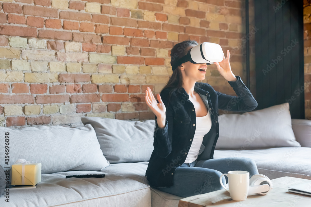 excited woman touching virtual reality headset while sitting on sofa - obrazy, fototapety, plakaty 