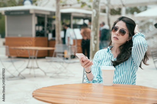 portrait of beautiful young asian chinese woman reading text message on mobile phone at coffee shop outdoor. lady in sunglasses enjoy sunshine sitting at table with takeaway drink in disposable cup