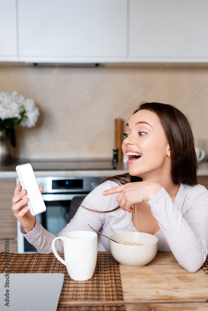 excited girl pointing with finger at smartphone with white screen near cup and bowl excited girl pointing with finger at smartphone with white screen near cup and bowl