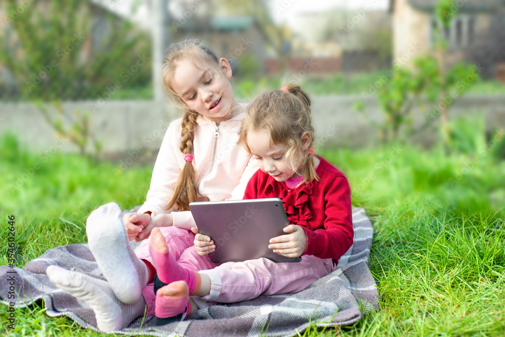 Smiling children girls sit on a blanket at the garden or park and playing with digital tablet together. Smiling children girls sit on a blanket at the garden or park and playing with digital tablet together.