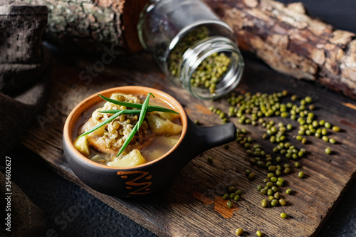 Soup with mash and potatoes. Decorated with green onions and peas of small beans from a jar. Serving in a ceramic pot on a charred wooden background with fabric and natural driftwood