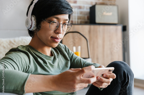 Image of asian man wearing headphones using cellphone in apartment