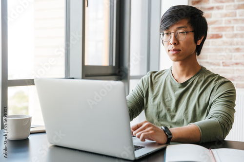 Image of handsome young asian man using laptop in apartment