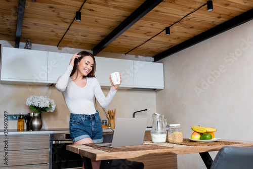 happy freelancer holding cup near laptop, jug with milk and fruits in kitchen