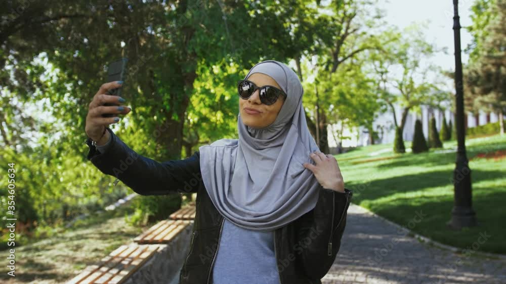 Muslim woman in casual outfit, sunglasses and hijab. She is smiling, showing victory sign, taking selfie on her smartphone, posing in park. Сlose up Muslim woman in casual outfit, sunglasses and hijab. She is smiling, showing victory sign, taking selfie on her smartphone, posing in park. Сlose up