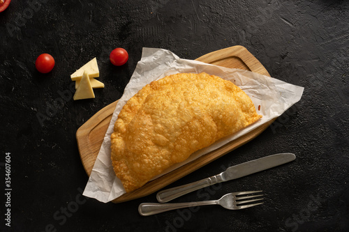 Cheburek with meat, cheese and tomatoes on a wooden board next to cutlery, and ingredients on a black table. Top view.