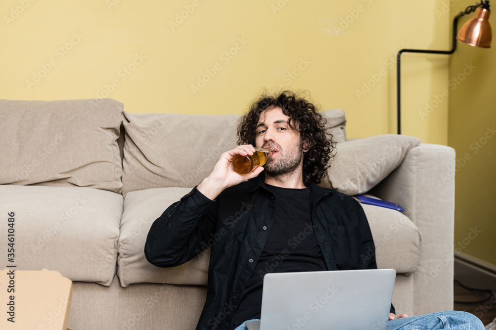 Fototapeta premium Handsome man looking at camera while drinking beer near laptop and pizza box at home