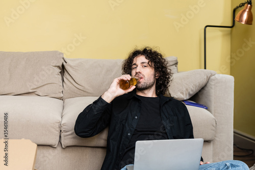 Handsome man looking at camera while drinking beer near laptop and pizza box at home