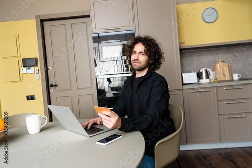 handsome freelancer smiling at camera while holding credit card and typing on laptop near smartphone with blank screen