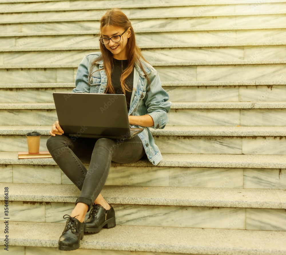 Young modern woman student in a denim jacket sitting on the stairs and watching video on laptop. Distance learning. Modern youth concept. Young modern woman student in a denim jacket sitting on the stairs and watching video on laptop. Distance learning. Modern youth concept.