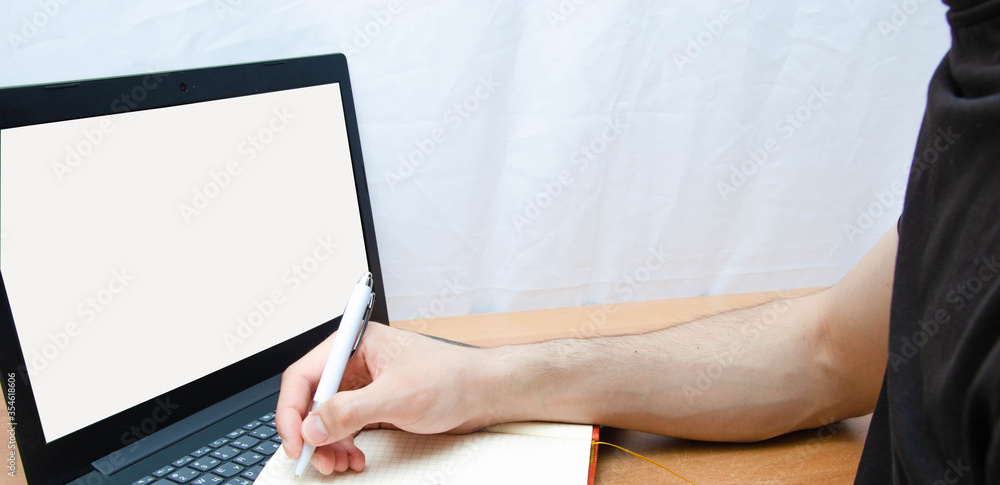 Naklejka premium A man at a laptop. A guy in a black T-shirt sits at a table and works on a laptop, typing. Writes notes, plans, tasks with a pen in a notebook. White screen laptop