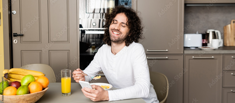 panoramic shot of smiling man chatting on smartphone during breakfast in kitchen